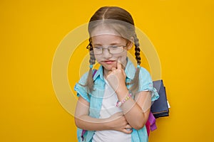 Back to school. Beautiful little girl in glasses and with pigtails with books in a school backpack thoughtfully holds a finger