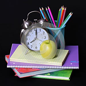 Back to school concept. An apple, colored pencils and alarm clock on pile of books over black