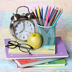 Back to school concept. apple, colored pencils, glasses and alarm clock on pile of books over the map