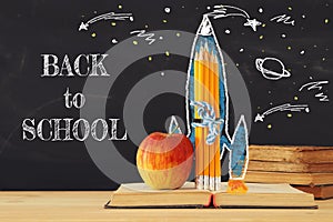 Back to school concept. rocket sketch and pencils over open book in front of classroom blackboard.