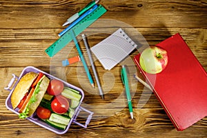 Back to school concept. School supplies, books, apple and lunch box with burgers and fresh vegetables on wooden table. Top view