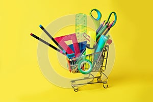 Back to school concept with shopping cart and colorful pencils, square ruler, scissors, clips, markers on pastel yellow backdrop.