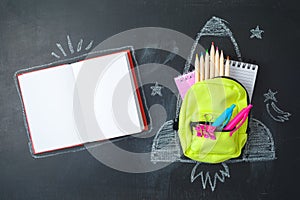 Back to school concept with small bag backpack, school supplies and rocket sketch over chalkboad background. Top view from above
