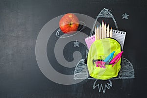 Back to school concept with small bag backpack, school supplies and rocket sketch over chalkboard background. Top view from above