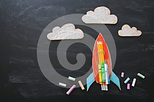 Back to school concept. Top view image of handmade cardboard rocket and clouds with pencils over classroom blackboard background