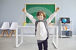 Back to school. Funny schoolboy with backpack raised his hands up while standing in class.