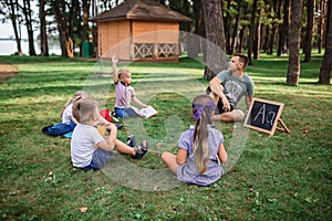 Back to school. Kindergarten and elementary scholars sitting with teacher on grass at open-air class