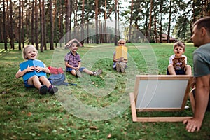 Back to school. Kindergarten and elementary scholars sitting with teacher on grass at open-air class