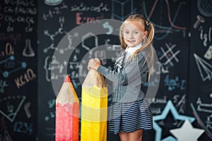 Back to school! A little blonde girl in school uniform stands with two very large red and yellow pencils on the floore against