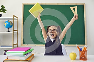 Back to school. Little happy schoolchild raised hands up against the background of a green blackboard in a classroom.