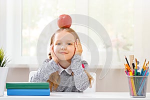 Back to school. Little student girl sitting at a table with a red apple on her head.