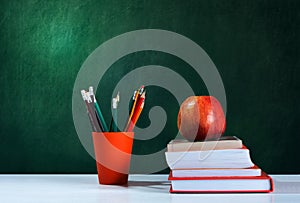 Back to school, orange pencil holder, stack of books on white table with red apple, empty green school board background, education