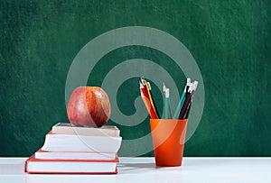 Back to school, orange pencil holder, stack of books on white table with red apple, empty green school board background, education