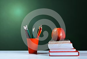 Back to school, orange pencil holder, stack of books on white table with red apple, empty green school board background, education