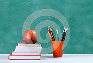 Back to school, orange pencil holder, stack of books on white table with red apple, empty green school board background, education