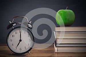 Back to school, vintage old alarm clock near books stacked against blackboard and apple fruit