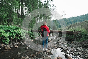 Back of tourist woman in red raincoat stands in forest on rocks in mountain stream, looking forward. Girl walking in mountainous