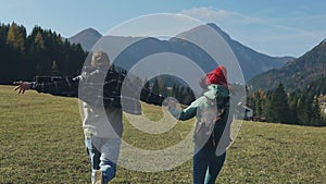 Back of two guys holding hands with backpacks on summer vacation or holidays running through alpine meadows. Silhouette