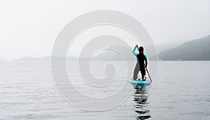 Back view active sportswoman in diving suit floating on stand up paddle at fog sea scenery