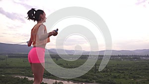 Back view on active young brunette woman jumping with skipping rope on the mountain peak. Green mountain ladscape