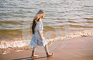 Back view of little girl with long hair in white dress walking on tropical beach vacation