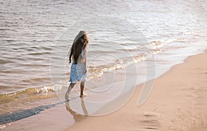 Back view of little girl with long hair in white dress walking on tropical beach vacation