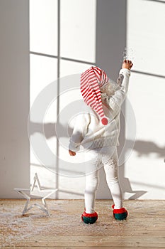 Back view of adorable small kid elf wears white costume, santa s hat and elf s shoes, plays with artificial snow, stands