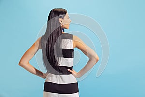 Back view of adorable young woman in summer striped dress posing isolated on blue background. Concept of beauty, art