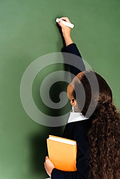 back view of african american schoolgirl writing on chalkboard while holding book.