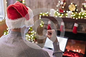 Back view of angry man freelancer in santa claus hat, raising and swinging fist on laptop screen sitting near christmas tree