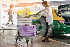 Back view of Asian female passenger with big roller luggage stand to wait for the car to pick up at airport departure terminal.