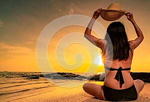 Back view of Asian plump woman wear bikini and hand holding straw hat sit on sand beach relax and enjoy holiday at tropical beach