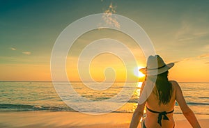 Back view of Asian woman wear bikini and straw hat sit on sand beach relax and enjoy holiday at tropical paradise beach. Woman