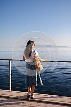 Back view of attractive woman in denim style stand in front of sea landscape. Happy woman looking on sunset