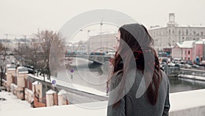 Back view. Attractive young brunette girl standing on the bridge and looks at the snow-covered winter town.