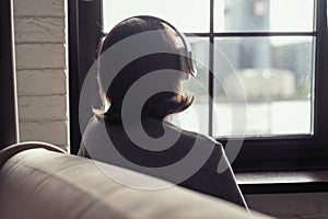 Back view of Ñaucasian young woman in headphones enjoying listening music sitting near window of apartments.