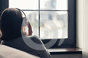 Back view of Ñaucasian young woman in headphones enjoying listening music sitting near window of apartments.