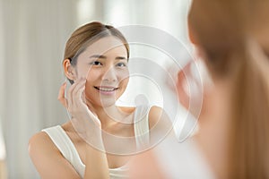 Back view of beautiful Asian young woman looking in the mirror after wake up preparing skin for make up. Healthy female looking