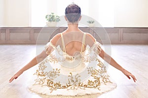 Back view of beautiful ballerina in cream dress sitting and warms up her hands on wooden floor in ballet studio