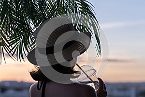 Back view of a beautiful girl with cold water in a glass in a straw hat against the background of the sea in branches of palm tree