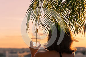 Back view of a beautiful girl with lemon water in a plastic glass against the background of the sea in branches of palm trees.