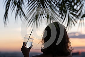 Back view of a beautiful girl with mineral water in a glass against the background of the sea in branches of palm trees. Sunset