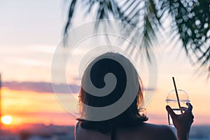 Back view of a beautiful girl with mineral water in a glass against the background of the sea in branches of palm trees. Sunset