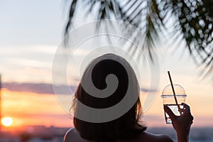 Back view of a beautiful girl with mineral water in a glass against the background of the sea in branches of palm trees. Sunset