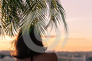 Back view of a beautiful girl in a straw hat against the background of the sea in branches of palm trees. Sunset beach. Summer