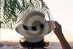 Back view of a beautiful girl in a straw hat against the background of the sea in branches of palm trees. Sunset beach. Summer