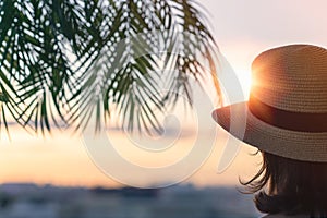 Back view of a beautiful girl in a straw hat against the background of the sea in branches of palm trees. Sunset beach. Summer