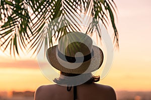 Back view of a beautiful girl in a straw hat against the background of the sea in branches of palm trees. Sunset beach. Summer