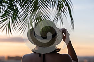 Back view of a beautiful girl in a straw hat against the background of the sea in branches of palm trees. Sunset beach. Summer