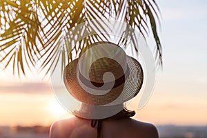 Back view of a beautiful girl in a straw hat against the background of the sea in branches of palm trees. Sunset beach. Summer